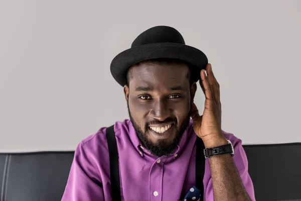 Портрет улыбающегося африканского стильного человека в шляпе, сидящего на черном диване, изолированном на сером — стоковое фото