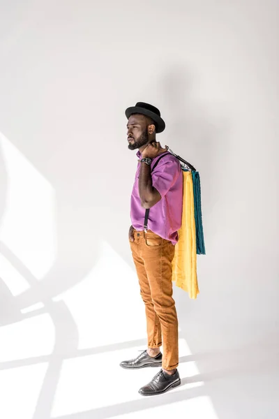 Jeune homme afro-américain en vêtements à la mode et cintres tenant chapeau avec textile coloré sur fond gris — Photo de stock