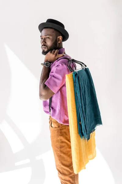 Side view of african american man in fashionable clothing and hat holding hangers with colorful textile on grey background — Stock Photo