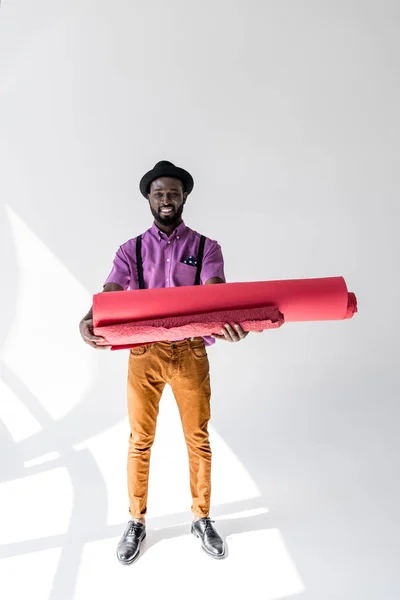 Smiling african american in hat holding pink wrapping paper rolls in hands on grey backdrop — Stock Photo
