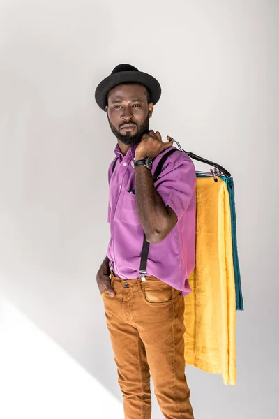 Retrato de hombre afroamericano en ropa de moda y sombrero sosteniendo perchas con textiles de colores sobre fondo gris - foto de stock