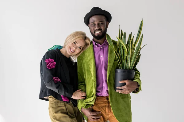 Портрет улыбающейся межрасовой стильной пары с зеленым растением в цветочном горшке, позирующем изолированно на сером — стоковое фото