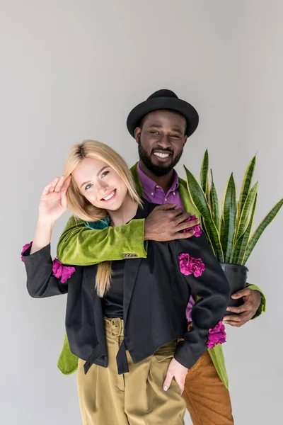 Porträt eines lächelnden gemischtrassigen stilvollen Paares mit grüner Pflanze im Blumentopf, das isoliert auf grau posiert — Stockfoto