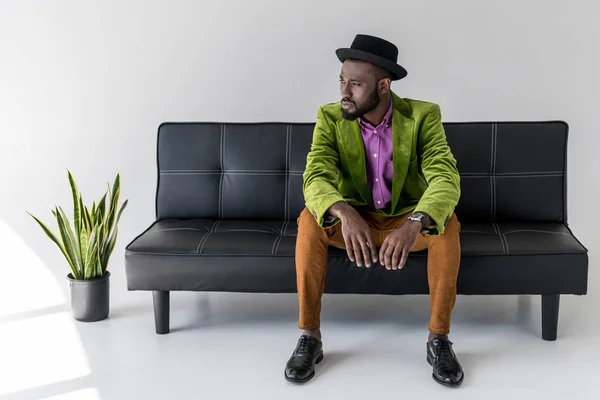 Hombre de moda afroamericano pensativo en sombrero sentado en sofá negro - foto de stock