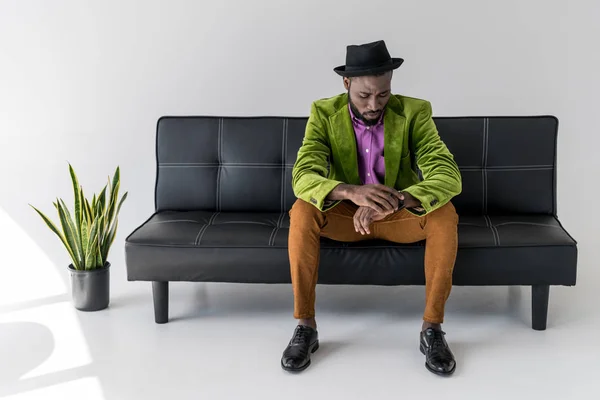 Afro americano elegante homem em chapéu verificando o tempo enquanto sentado no sofá preto — Fotografia de Stock