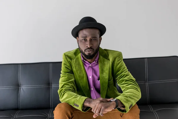Portrait of african american fashionable man in hat sitting on black sofa pkisolated on grey — Stock Photo