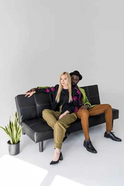 Мультикультурная модная пара отдыхает на черном диване на сером фоне — стоковое фото