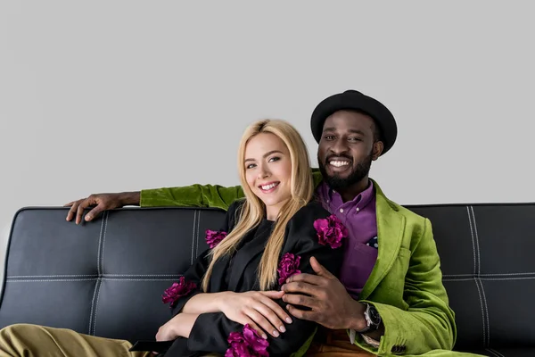 Porträt eines lächelnden multikulturell modischen Paares, das sich auf einem schwarzen Sofa ausruht, isoliert auf grau — Stockfoto