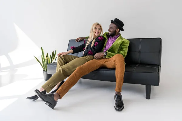 Веселая мультикультурная модная пара отдыхает на черном диване на сером фоне — стоковое фото