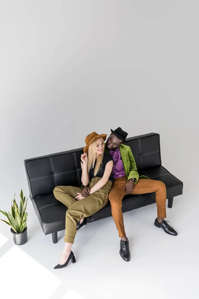 Высокий угол обзора улыбающейся мультикультурной модной пары, сидящей на черном диване — стоковое фото