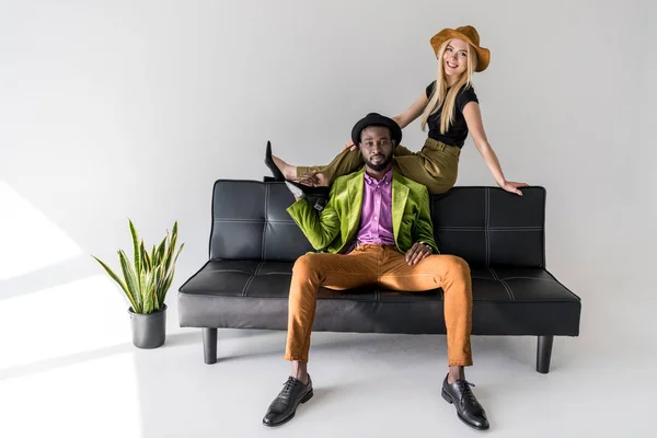 Мультикультурная модная пара в шляпах на черном диване на сером фоне — стоковое фото