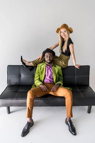 Junges multikulturelles Modepaar in Hüten auf schwarzem Sofa isoliert auf grau — Stockfoto