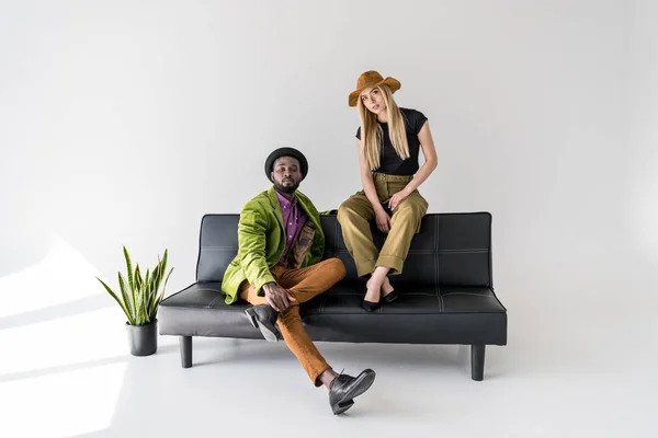 Young multicultural fashionable couple sitting on black sofa on grey backdrop — Stock Photo