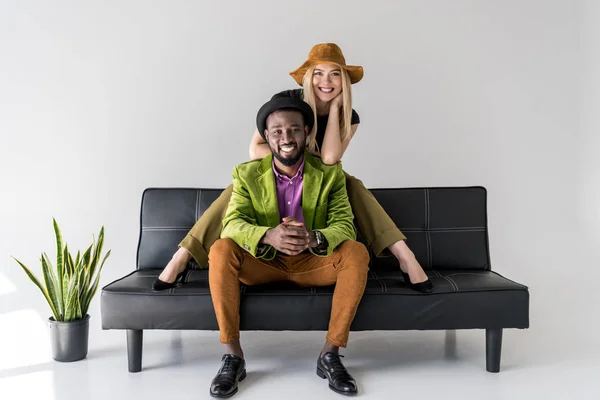 Cheerful multicultural fashionable couple in hats sitting on black sofa on grey background — Stock Photo