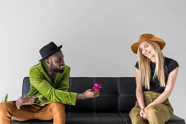 African american fashionable man presenting flower to caucasian girlfriend on black sofa isolated on grey — Stock Photo