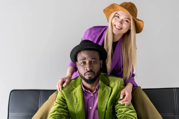 Retrato de atractiva pareja multicultural de moda en sombreros sentados en sofá negro aislado en gris - foto de stock