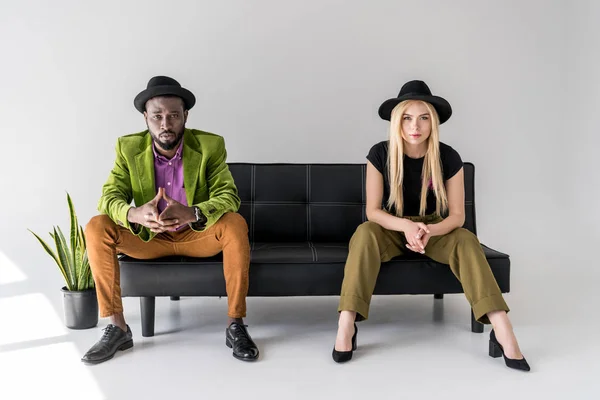 Pareja multicultural de moda en sombreros sentados en sofá negro sobre fondo gris - foto de stock