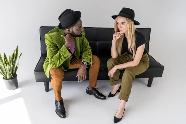 Multicultural fashionable couple in hats looking at each other on black sofa on grey backdrop — Stock Photo