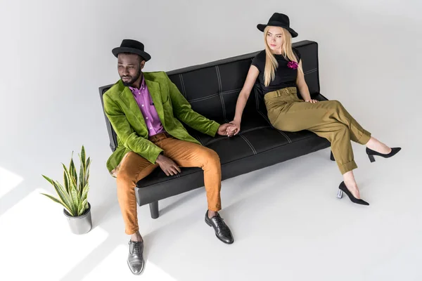 Pareja multicultural de moda en sombreros tomados de la mano en el sofá negro sobre fondo gris — Stock Photo