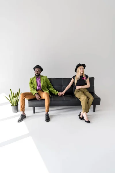 Joven pareja multiétnica cogida de la mano mientras está sentada en el sofá y mirando hacia otro lado en gris - foto de stock