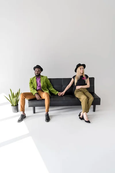 Young multiethnic couple holding hands while sitting on couch and looking away on grey — Stock Photo