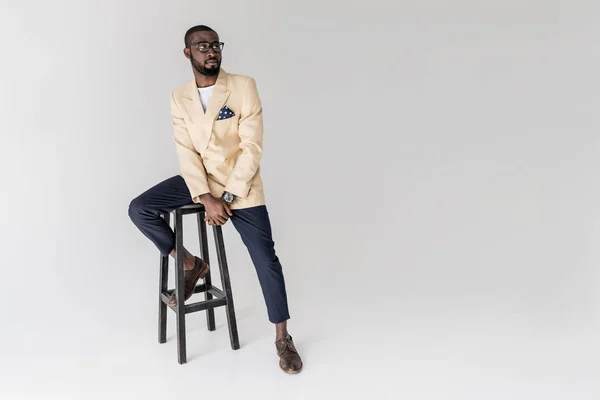 Vista completa del hombre afroamericano con estilo en gafas sentadas en el taburete y mirando hacia otro lado aislado en gris - foto de stock