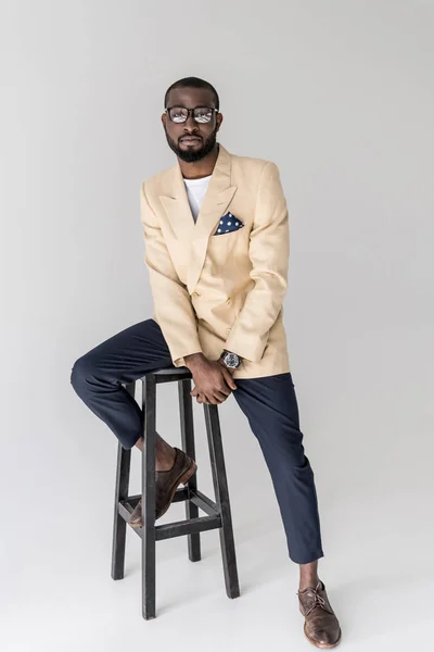 Красивий молодий стильний афроамериканський чоловік в окулярах сидить на стільці і дивиться на камеру на сірому — стокове фото