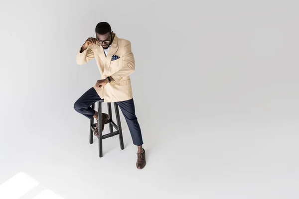 Високий кут зору модний афроамериканський чоловік сидить на табуреті і перевіряє наручний годинник ізольований на сірому — стокове фото