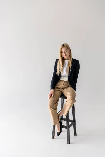 Full length view of beautiful stylish blonde girl sitting on chair and looking at camera isolated on grey — Stock Photo