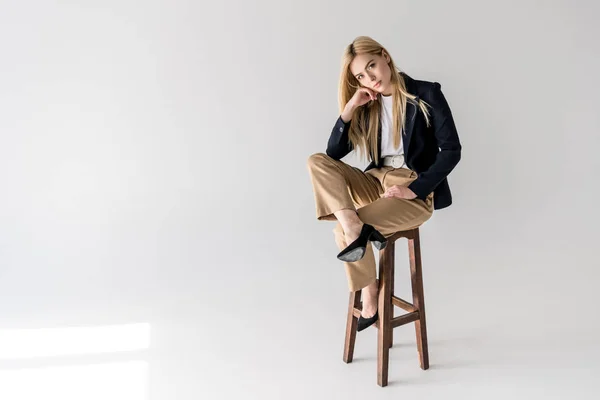 Attractive young blonde woman in stylish clothes sitting on stool and looking at camera on grey — Stock Photo