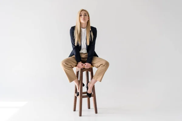 Beautiful young blonde woman in stylish clothes sitting on stool and looking at camera isolated on grey — Stock Photo
