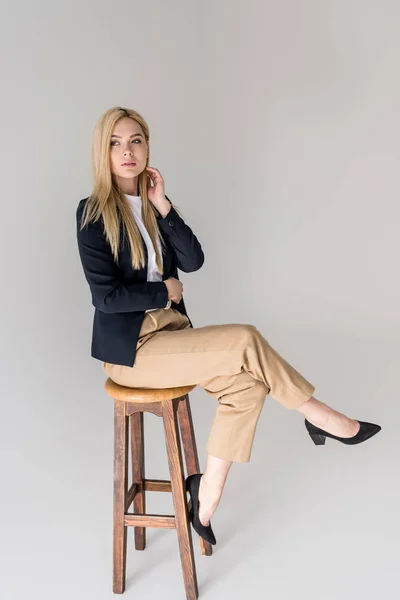 Vue pleine longueur de belle fille blonde élégante assise sur un tabouret isolé sur du gris — Photo de stock