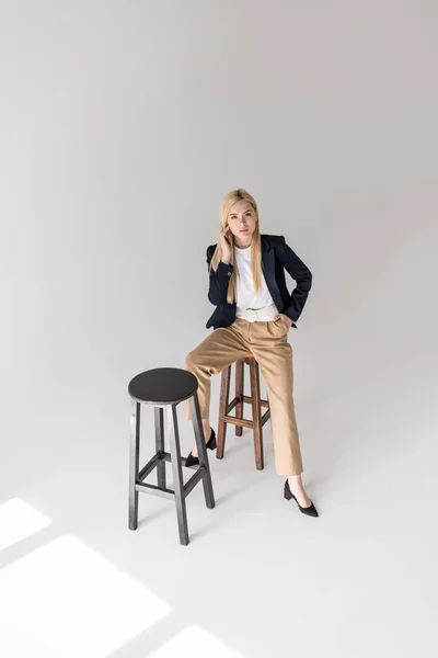 High angle view of beautiful stylish blonde girl sitting on stool and looking at camera on grey — Stock Photo