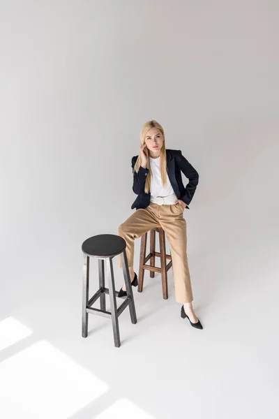 Vue grand angle de belle fille blonde élégante assise sur un tabouret et regardant la caméra sur gris — Photo de stock