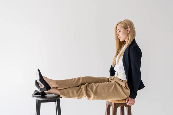 Vue latérale de belle fille blonde à la mode assise sur un tabouret et regardant les jambes isolées sur du gris — Photo de stock