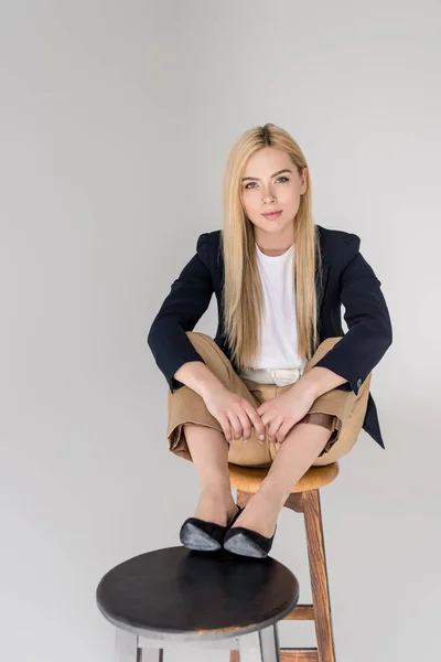 Bella ragazza bionda elegante seduta su due sedie e sorridente alla fotocamera isolata sul grigio — Foto stock