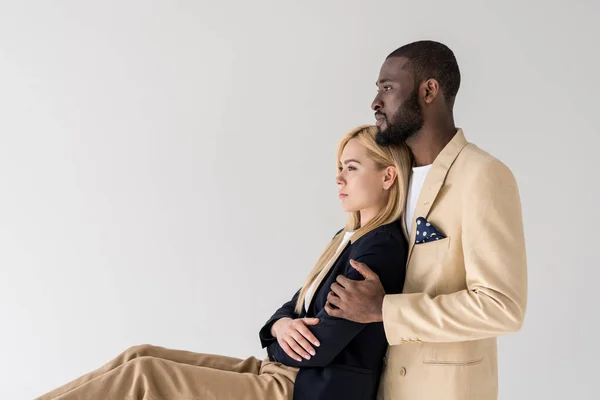 Vue de côté de jeune couple multiethnique à la mode embrassant et regardant loin isolé sur gris — Photo de stock