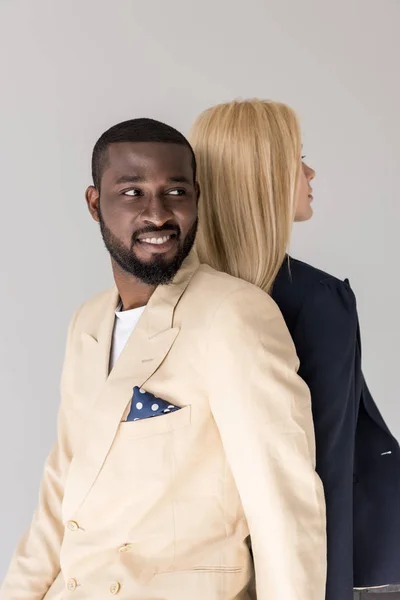 Lächelnd hübscher junger afrikanisch-amerikanischer Mann, der wegschaut, während er Rücken an Rücken mit seiner Freundin in grau isoliert sitzt — Stockfoto