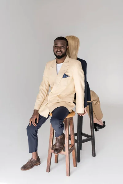 Elegante jovem casal multiétnico sentado de volta para trás, homem sorrindo para a câmera isolada em cinza — Fotografia de Stock