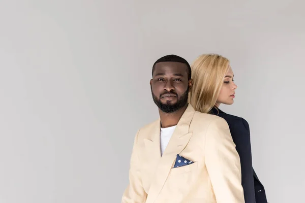 Stylish young multiethnic couple sitting back to back, man looking at camera isolated on grey — Stock Photo