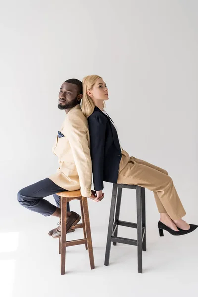 Vista lateral de la elegante pareja joven multiétnica sentada espalda con espalda y cogida de la mano en gris - foto de stock