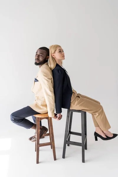 Side view of stylish young multiethnic couple sitting back to back and holding hands on grey — Stock Photo