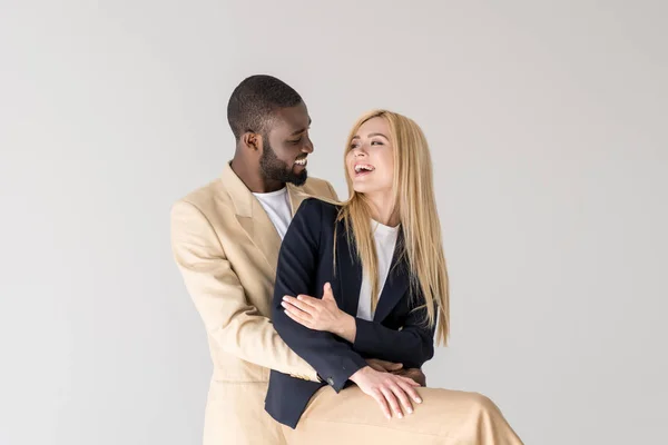 Happy stylish young multiethnic couple hugging and smiling each other isolated on grey — Stock Photo