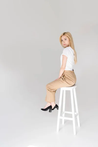 Beautiful young blonde woman sitting on stool and looking at camera isolated on grey — Stock Photo