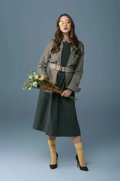 Красивая модная девушка позирует в осеннем наряде с букетом цветов, изолированных на сером — стоковое фото