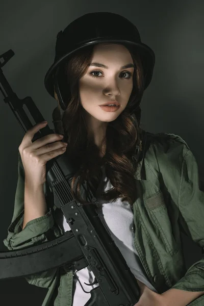 Beautiful caucasian young girl in military helmet with rifle, on grey with shadows — Stock Photo