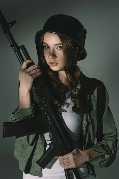 Attractive young girl in military helmet with rifle, on grey with shadows — Stock Photo