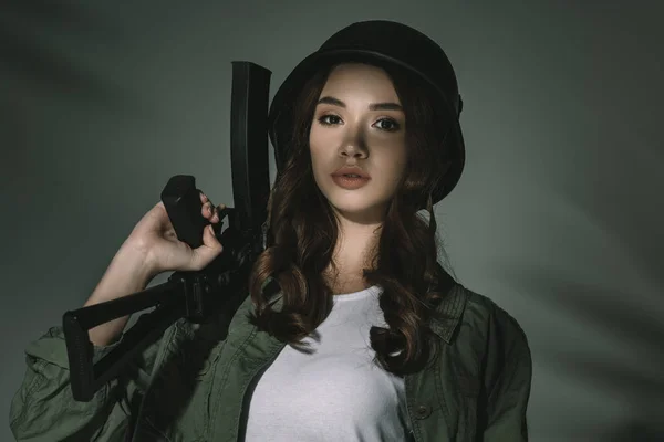 Attractive girl in military helmet with rifle, on grey with shadows — Stock Photo