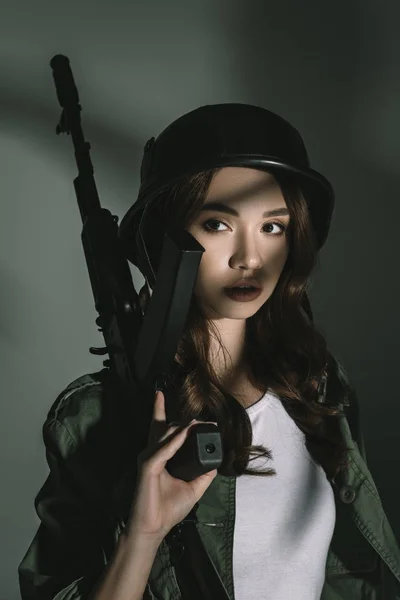 Beautiful girl posing in military helmet with rifle, on grey with shadows — Stock Photo