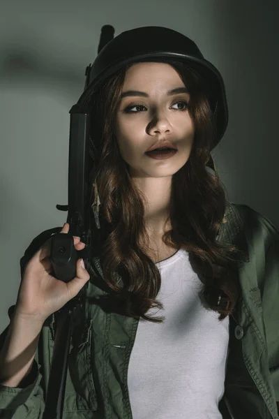 Hermoso soldado posando en casco militar con rifle, en gris con sombras - foto de stock